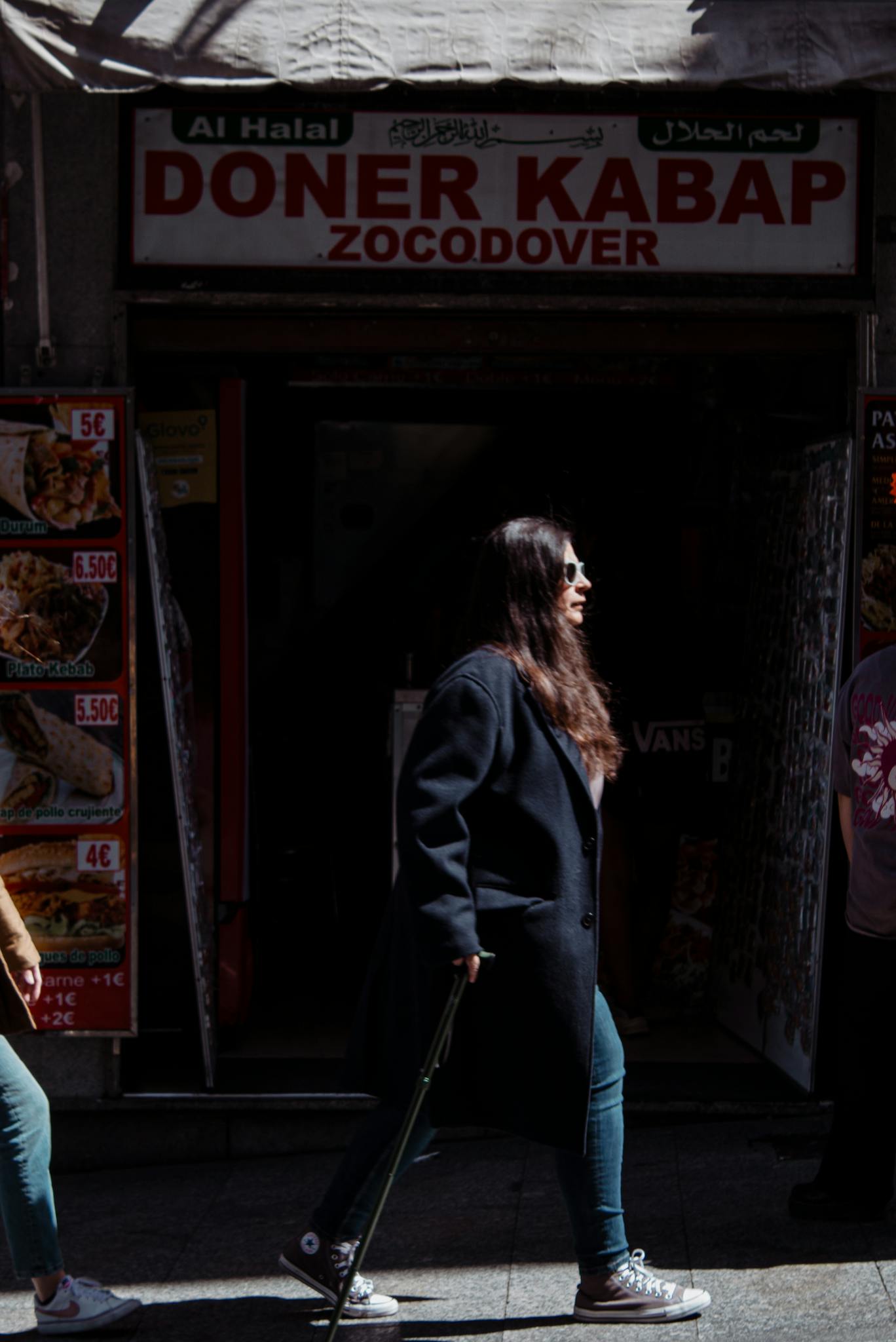 A woman in a coat and sunglasses walks past a doner kebab shop on a city sidewalk.